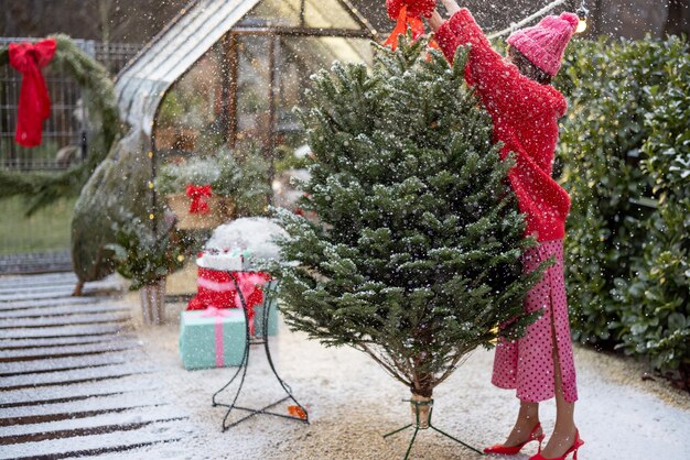 Young woman in red decorates lush Christmas tree with festive bows at backyard of her house on snow fall preparing for a winter holidays