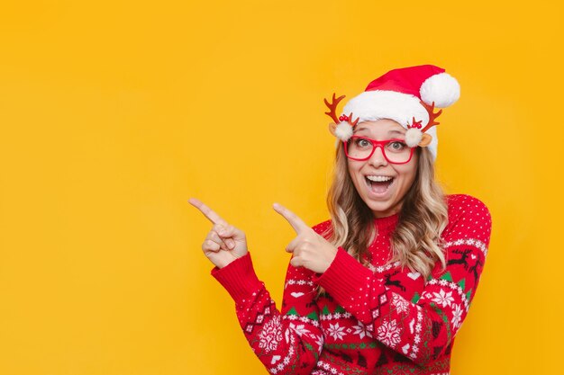 A young woman in a red deer sweater and Christmas Santa hat points at copy space for text or design