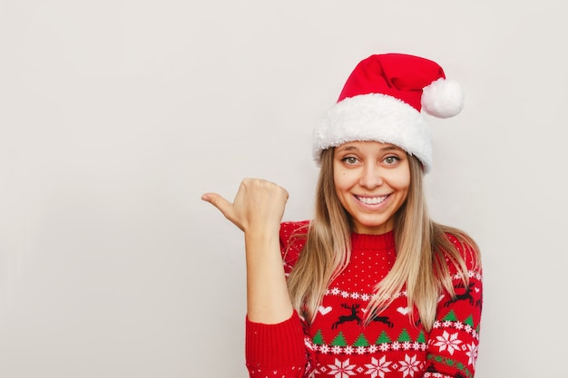 A young woman in a red deer sweater Santa hat points at empty copy space for text or design