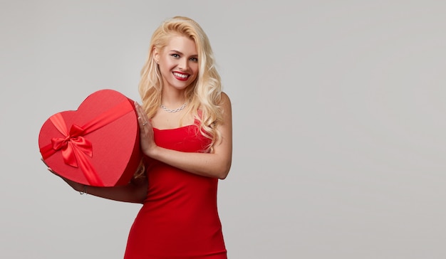 A young woman in a red dress blows a kiss. holds in her hands a heart shaped gift box. Valentines Day and March 8