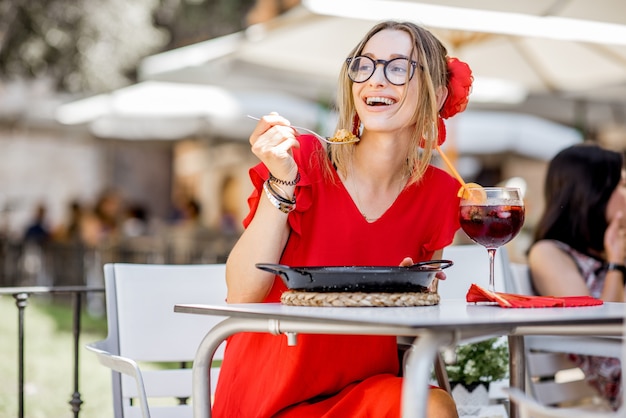 빨간 드레스를 입은 젊은 여성이 스페인 발렌시아에 있는 레스토랑에 야외에 앉아 전통적인 발렌시아 쌀 요리인 바다 빠에야를 먹고 있습니다.