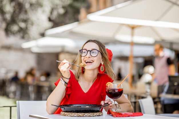 Young woman in red dress eating sea Paella, traditional Valencian rice dish, sitting outdoors at the restaurant in Valencia, Spain