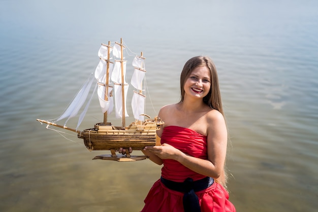 Young woman in red dress holding ship model on seashore