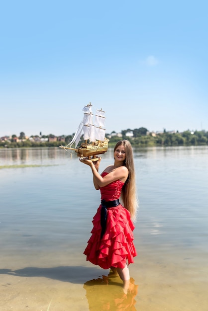 Young woman in red dress holding ship model on seashore