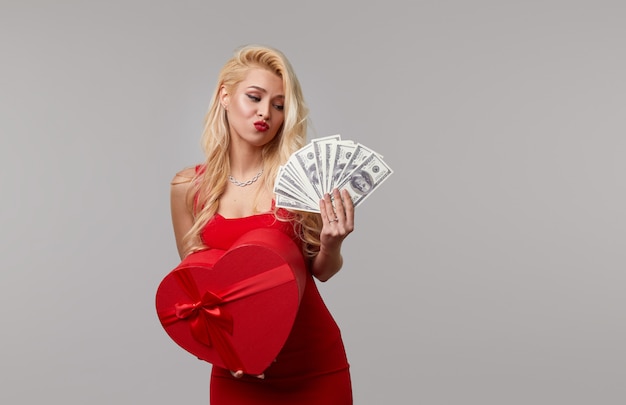 A young woman in a red dress holds in her hands cash banknotes money, dollars and a heart shaped gift box. Valentines Day and March 8