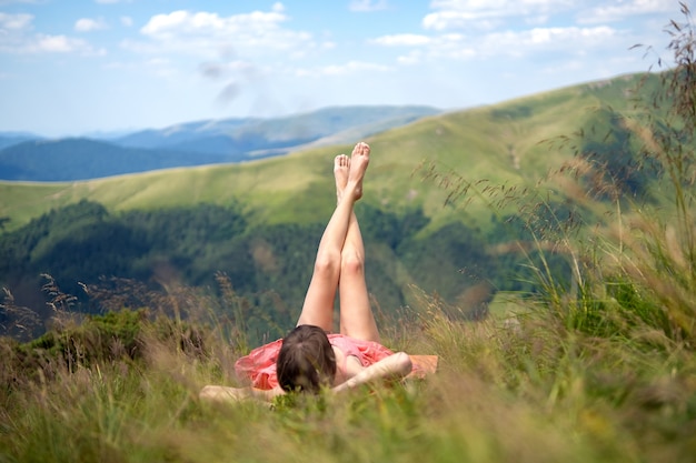 Giovane donna in abito rosso sdraiato sul verde campo erboso che riposa in una giornata di sole in montagna estate godendo della vista della natura.