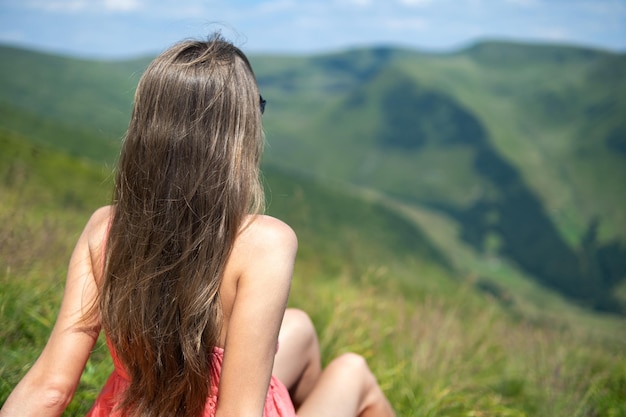 自然の景色を楽しむ夏の山々の晴れた日に緑の芝生のフィールドで休んで赤いドレスを着た若い女性。