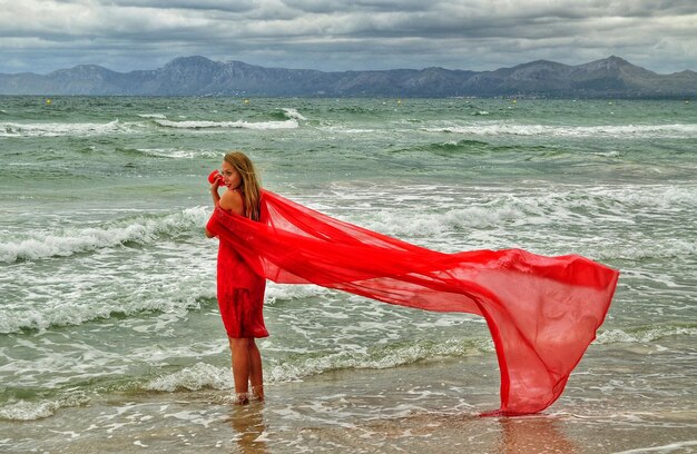 Photo young woman in red dress standing at beach against cloudy sky