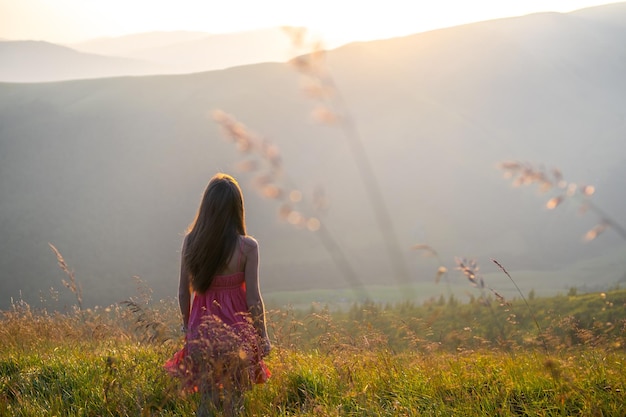 Young woman in red dress standing on grassy field on a windy evening in autumn mountains enjoying view of nature