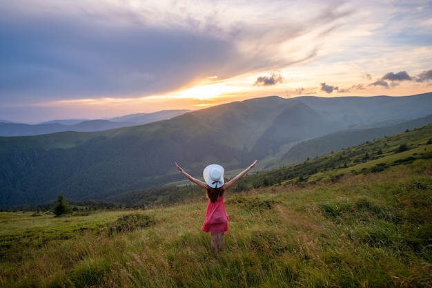 Young woman in red dress standing on grassy meadow on a windy evening in autumn mountains raising up her hands enjoying view of nature.