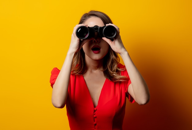 Young woman in red dress with binocular on yellow wall