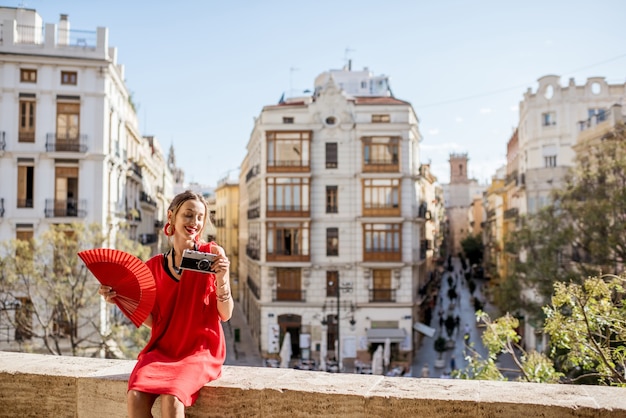 スペインの晴天時にバレンシアの街並みの美しい景色を楽しむ扇子と写真カメラと赤いドレスの若い女性