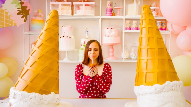 Young woman in red dress with short hair blowing out candle in muffin making wish congratulating herself on birthday