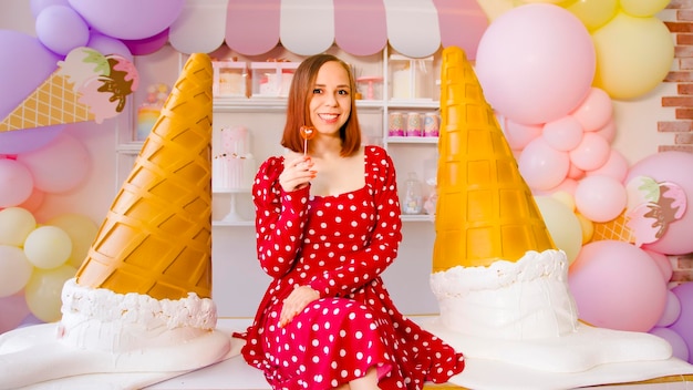 Young woman in red dress with short hair holding lollipop sitting on table in decorative confectionery