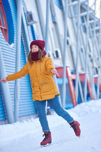 빨간 모자와 노란 재킷을 입은 젊은 여성이 서리가 내린 겨울날 경기장에서 아이스 스케이팅을 하고 있습니다. 수직 프레임.