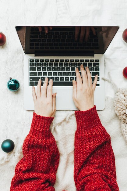 Giovane donna in maglione lavorato a maglia rosso che digita sul computer portatile sul letto bianco con coperta bianca decorata con palline rosse e blu di natale