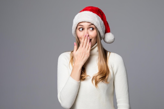 Young woman in a red Santa hat is isolated on a gray wall, an emotion of surprise and fright, covers her mouth with her hand