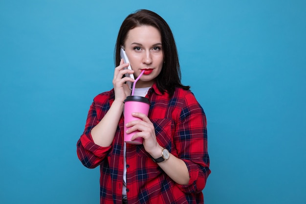 A young woman in a red shirt is talking on the phone and holding a glass of coffee