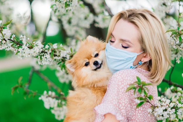 봄 날에 자연에 그녀의 얼굴에 의료 마스크와 젊은 여자와 빨간 스 피 츠. 코로나 바이러스 감염병 세계적 유행