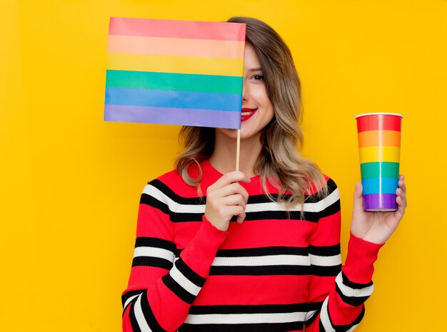 黄色のカップとLGBTの旗と赤い縞模様のセーターの若い女性