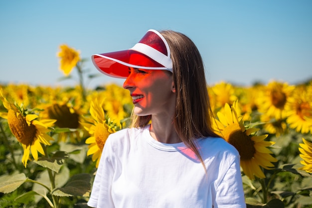 Giovane donna in visiera parasole rossa e maglietta bianca sul campo di girasoli.