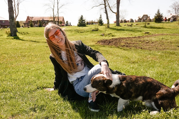 Young woman in red sunglasses plays with her corgi on a summer green lawn