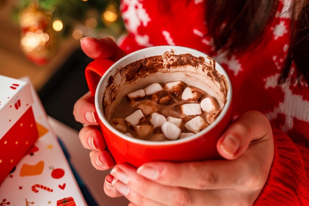 A young woman in a red sweater holds cocoa with marshmallows next to a Christmas tree