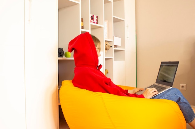 A young woman in a red sweater sits on an orange bean bag in the room and works at home with a laptop