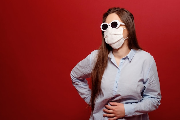 Young woman on red wall in protective medical mask and abdominal pain