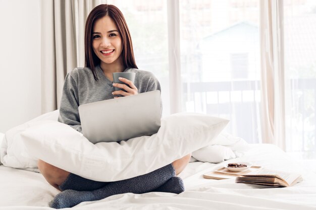 Giovane donna che si rilassa e che beve tazza di caffè o tè caldo facendo uso del computer portatile un giorno di inverno freddo nella camera da letto donna che controlla le app e lavoro sociali concetto di comunicazione e tecnologia