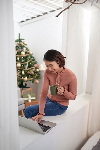 Young woman relaxing and drinking cup of hot coffee or tea using laptop computer on a cold winter day in the bedroom