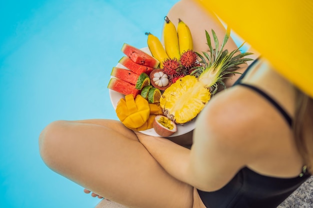 Young woman relaxing and eating fruit plate by the hotel pool Exotic summer diet Photo of legs with healthy food by the poolside top view from above Tropical beach lifestyle