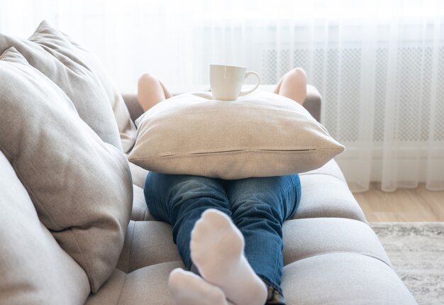 枕の上のコーヒーカップと彼女のソファでリラックスした若い女性