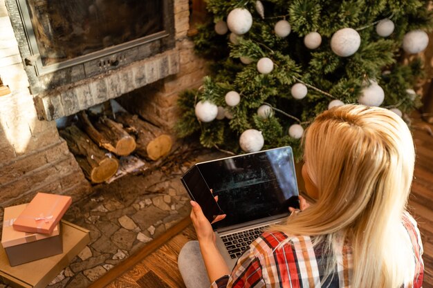 Giovane donna che si rilassa a casa durante il periodo natalizio seduta sul pavimento in soggiorno davanti all'albero di natale, navigando in internet sul suo computer portatile.