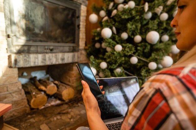 Young woman relaxing at home over Christmas sitting on the floor in the living room in front of the Xmas tree surfing the internet on her laptop computer.