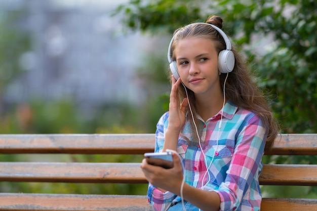 Young woman relaxing on an outdoor bench listening to music on stereo headphones looking to the side with a thoughtful expression