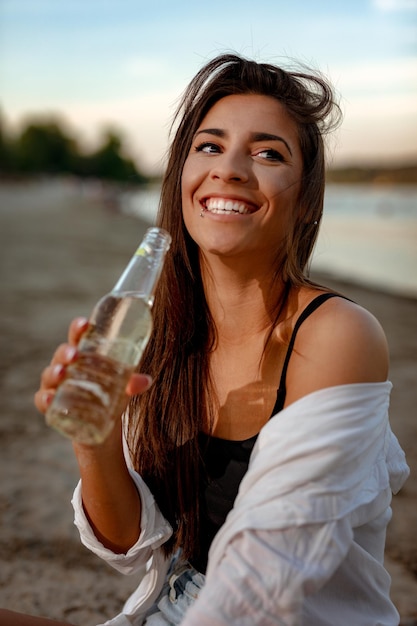 川岸で日没時にリラックスした若い女性。彼女は川のそばに座ってビールを飲んでいます。