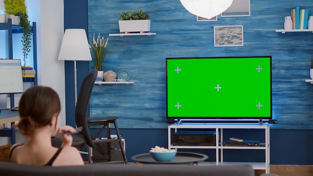 Young woman relaxing televison on green screen mockup display changing stations with remote control sitting comfortable on couch. Girl looking at favourite show on chroma key tv in living room.