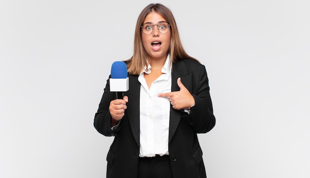 Young woman reporter feeling happy, surprised and proud, pointing to self with an excited, amazed look