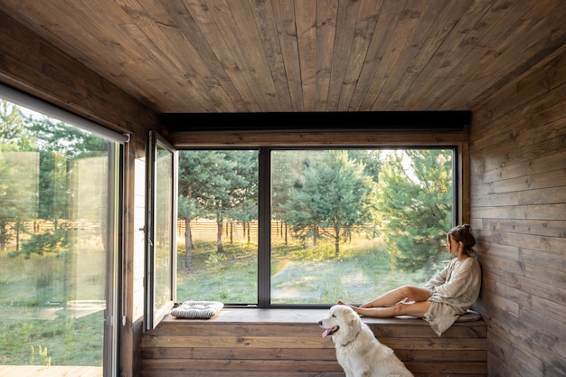 Young woman resting at beautiful country house or hotel, sitting on the window sill with pine forest view and big white dog sits near. Concept of solitude and recreation on nature with pet