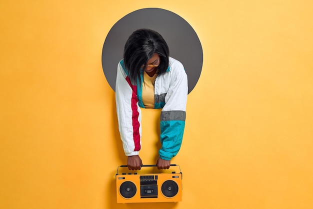 Young woman in retro jacket holding boombox in a circle hole in a bright wall copy space
