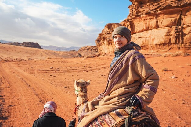 Young woman riding camel in Wadi Rum desert looking back over her shoulder smiling Its quite cold so she is wearing traditional Bedouin coat bisht and head scarf mountains far background