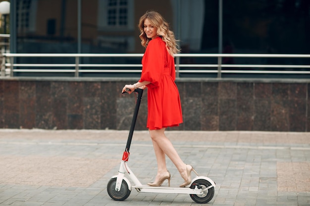 Young woman riding electric scooter in red dress at the city