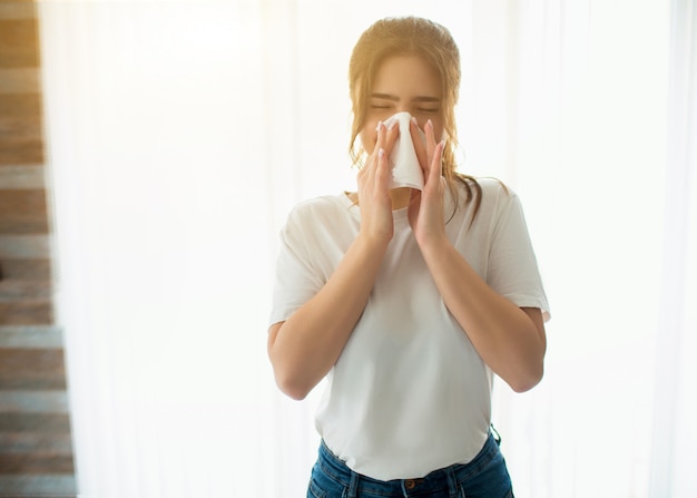 Young woman in room. Blowing her nose using white tissue. Sick ill person stand alone at window.