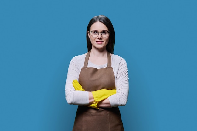 Young woman in rubber glove apron looking at camera with crossed arms on blue background Positive confident female cleaning service worker smiling housewife doing cleaning