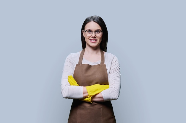 Young woman in rubber glove apron with crossed arms on grey background