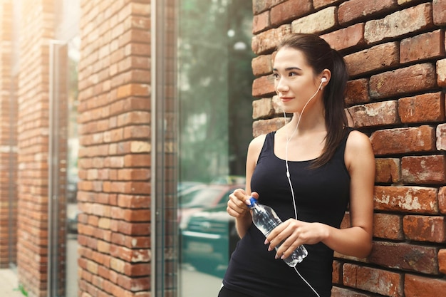 Young woman runner is having break, drinking water while jogging in city center, listening to music, brick wall background, copy space
