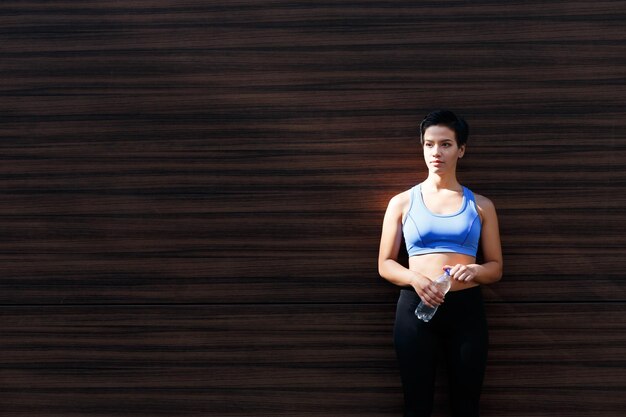 Young woman runner is having break, drinking water while jogging in city, leaning at dark wooden wall, copy space