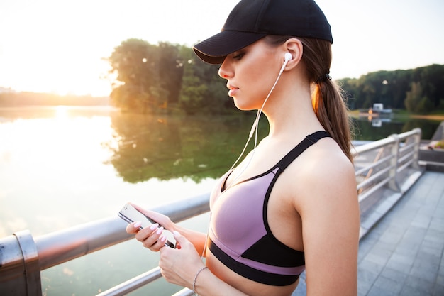 Young woman runner wearing armband and listening to music on earphones. Fit sportswoman taking a break from outdoors training