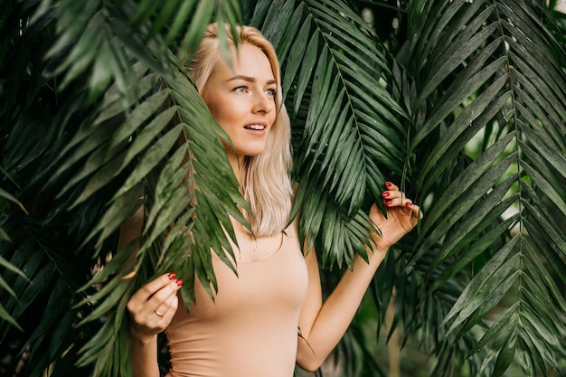 Young woman's face surrounded by tropical leaves. Closeup portrait. A cheerful smiling girl is surprised and looks away from copy space. Natural cosmetic, wellness, purity, skin care, beauty concept